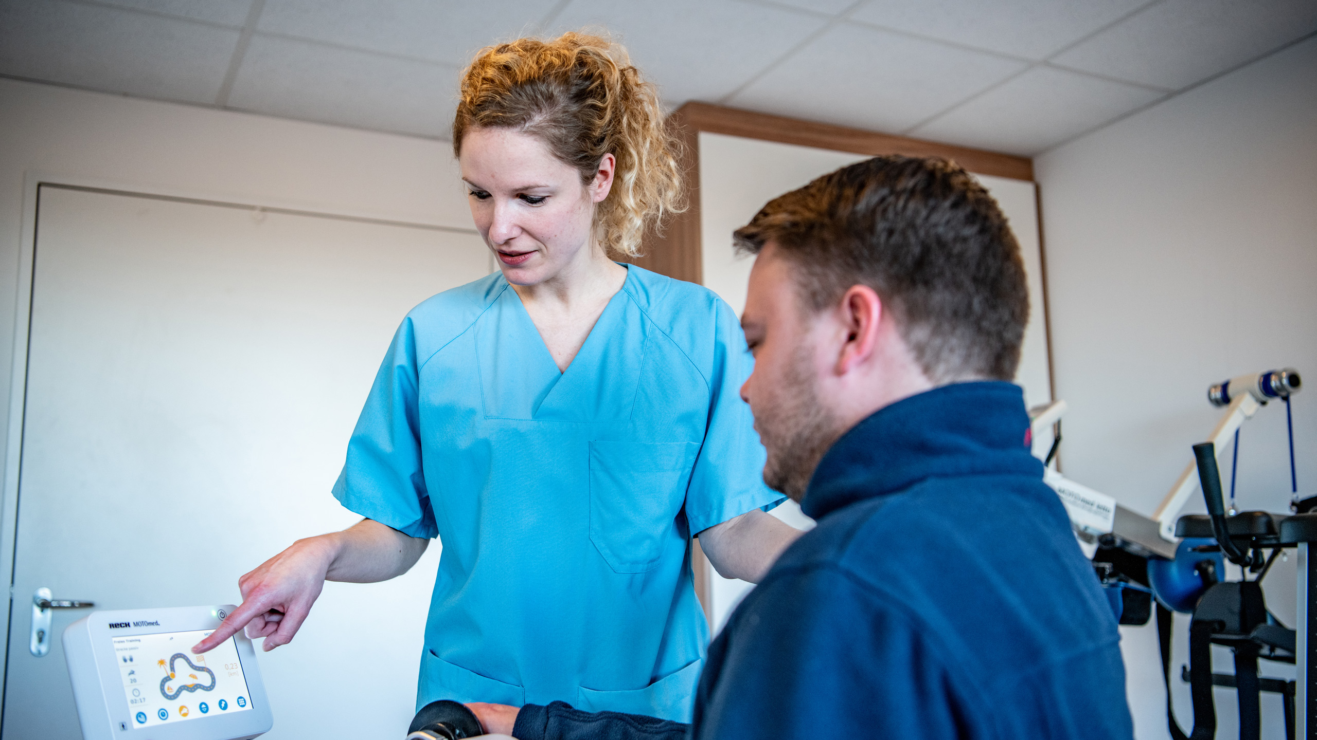 Mitarbeiterin der stationären Physiotherapie erklärt Patienten die Funktionen eines Gerätes